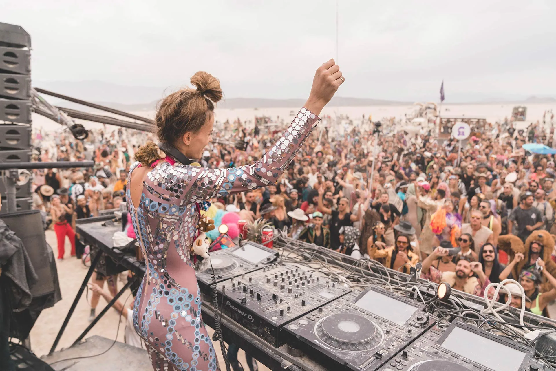 Deer Jade at the decks in a mirrored outfit, facing a crowd on the desert playa.