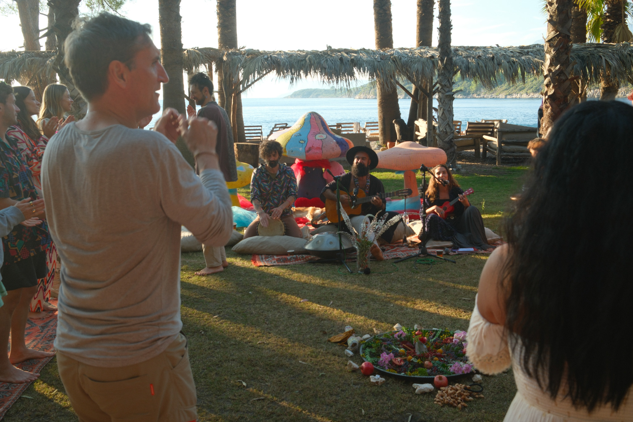 Musicians playing guitar, a small string instrument and hand drum beside the bay, with people gathered around them