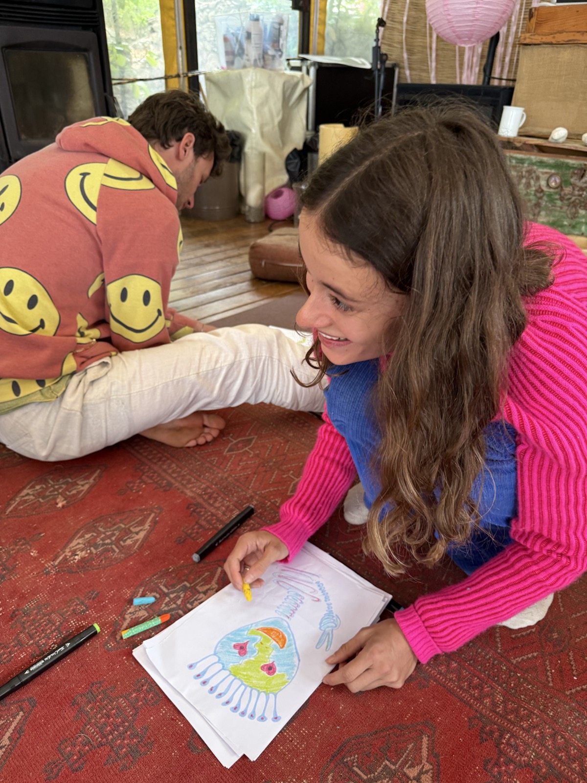 A participant in a pink sweater colours an alien drawing while seated on a carpet, with another participant drawing behind her