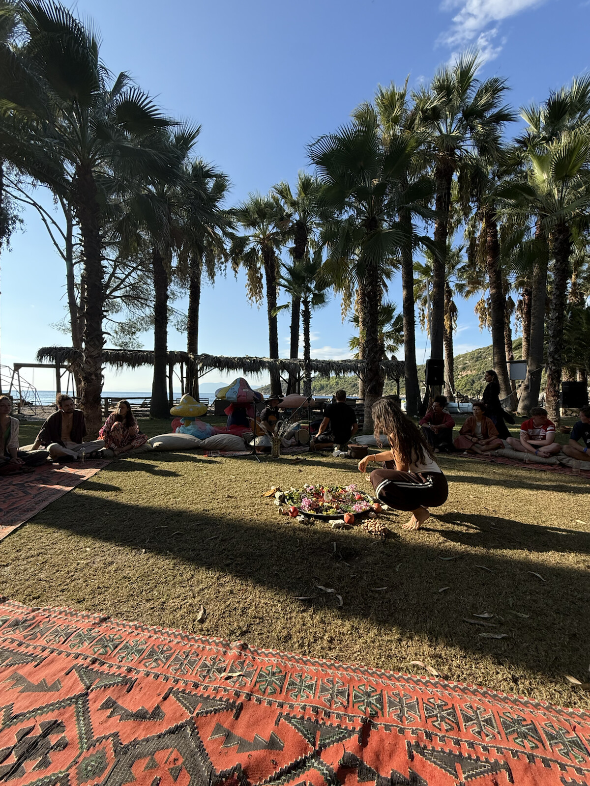 People resting beneath tall palms in the sunshine, with blue sky and the sea beyond